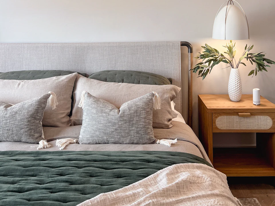 Linen headboard and layered bedding beside a timber nightstand, vase and pendant; illustrative interior, not a managed-property portfolio